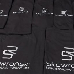 PATI-HAFT MARZENNA POŁETEK - Czarne koszulki z białym nadrukiem logo firmy budowlano-transportowej, ułożone obok siebie. Widoczne detale nadruku i materiału.