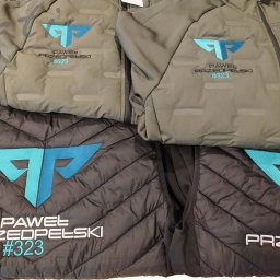 PATI-HAFT MARZENNA POŁETEK - Kurtki i kamizelki z haftowanym logo i nazwą 'Paweł Przedpełski #323' w odcieniach szarości i khaki. Widoczne detale zamków i pikowania.