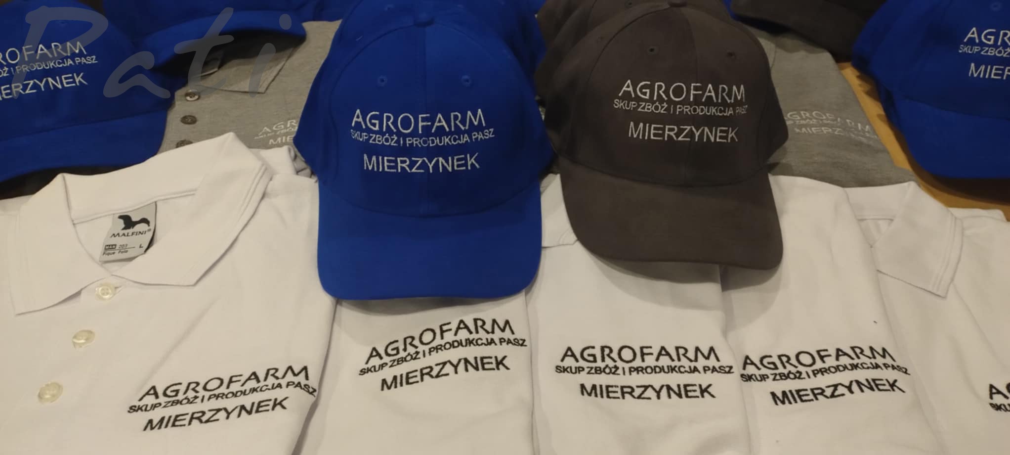 Haftowane logo 'AGROFARM' na białych koszulkach polo i czapkach z daszkiem w kolorze niebieskim i szarym. Prezentacja gotowych produktów.