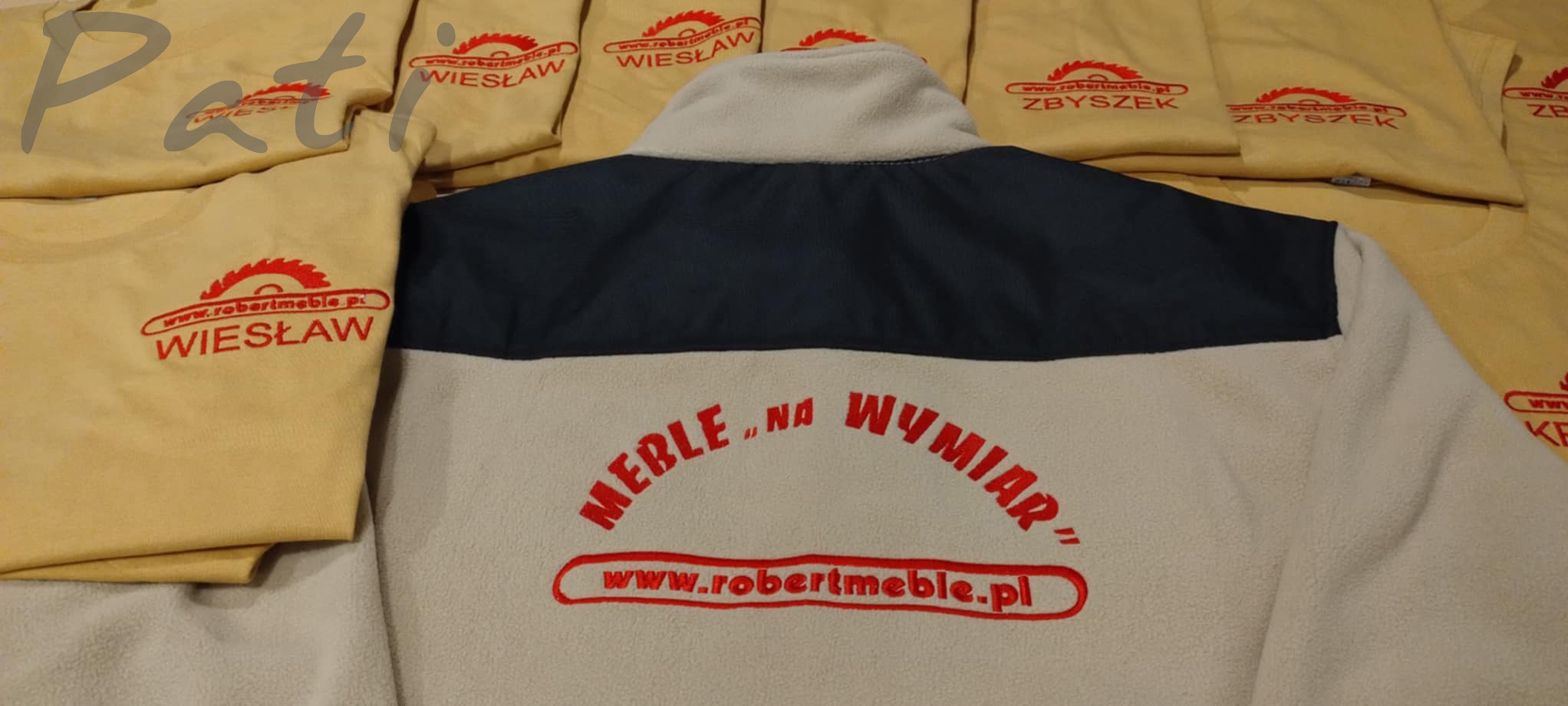 Beżowe koszulki i polar z haftowanym logo 'robertmeble.pl' i imionami (np. Wiesław, Zbyszek). Czerwony haft na odzieży. Widok z bliska, lekki kąt.