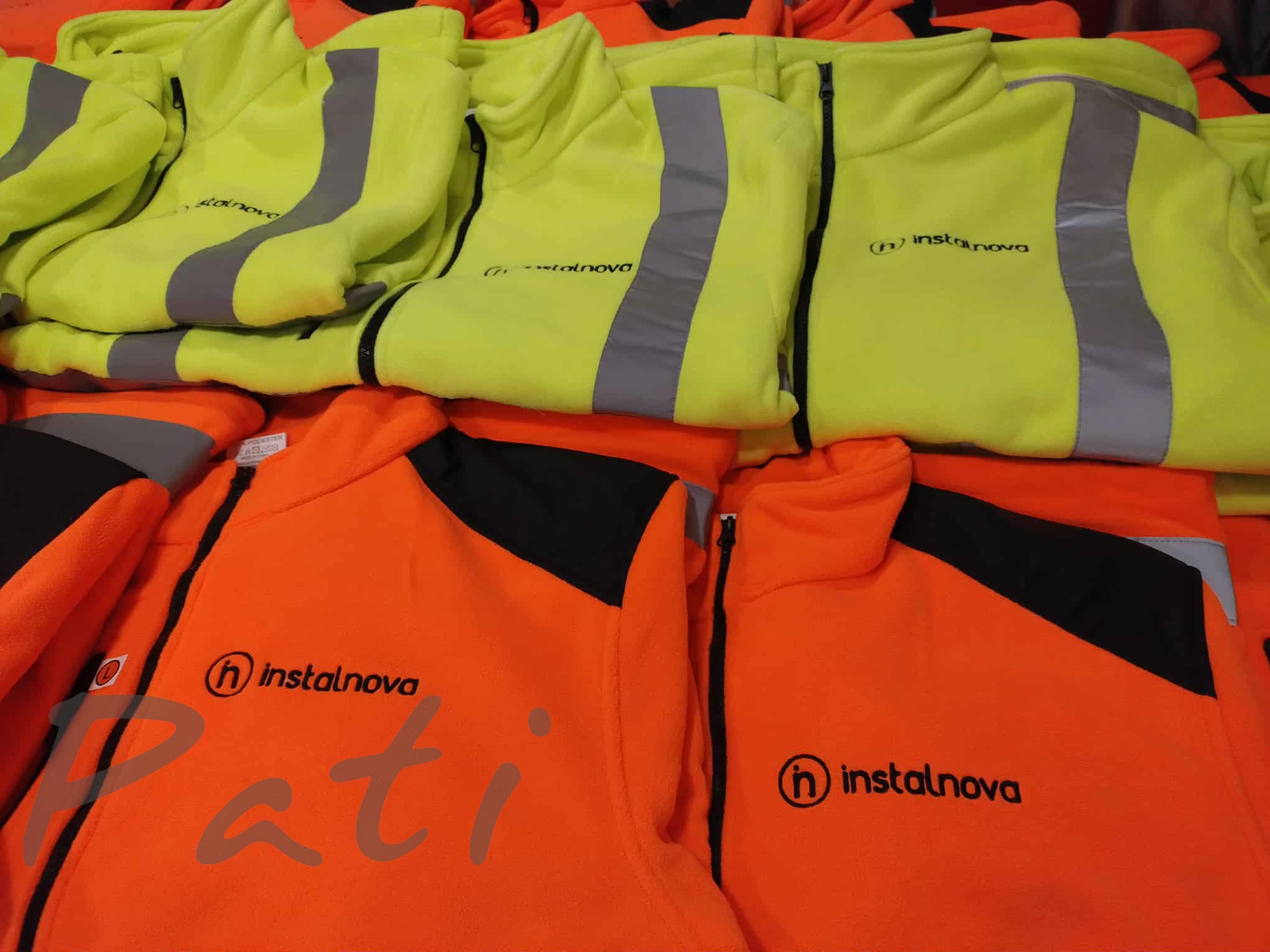 Stos kurtek polarowych w kolorach neonowej żółci i pomarańczy z odblaskowymi pasami i logo 'instalnova'. Widoczne detale haftu.