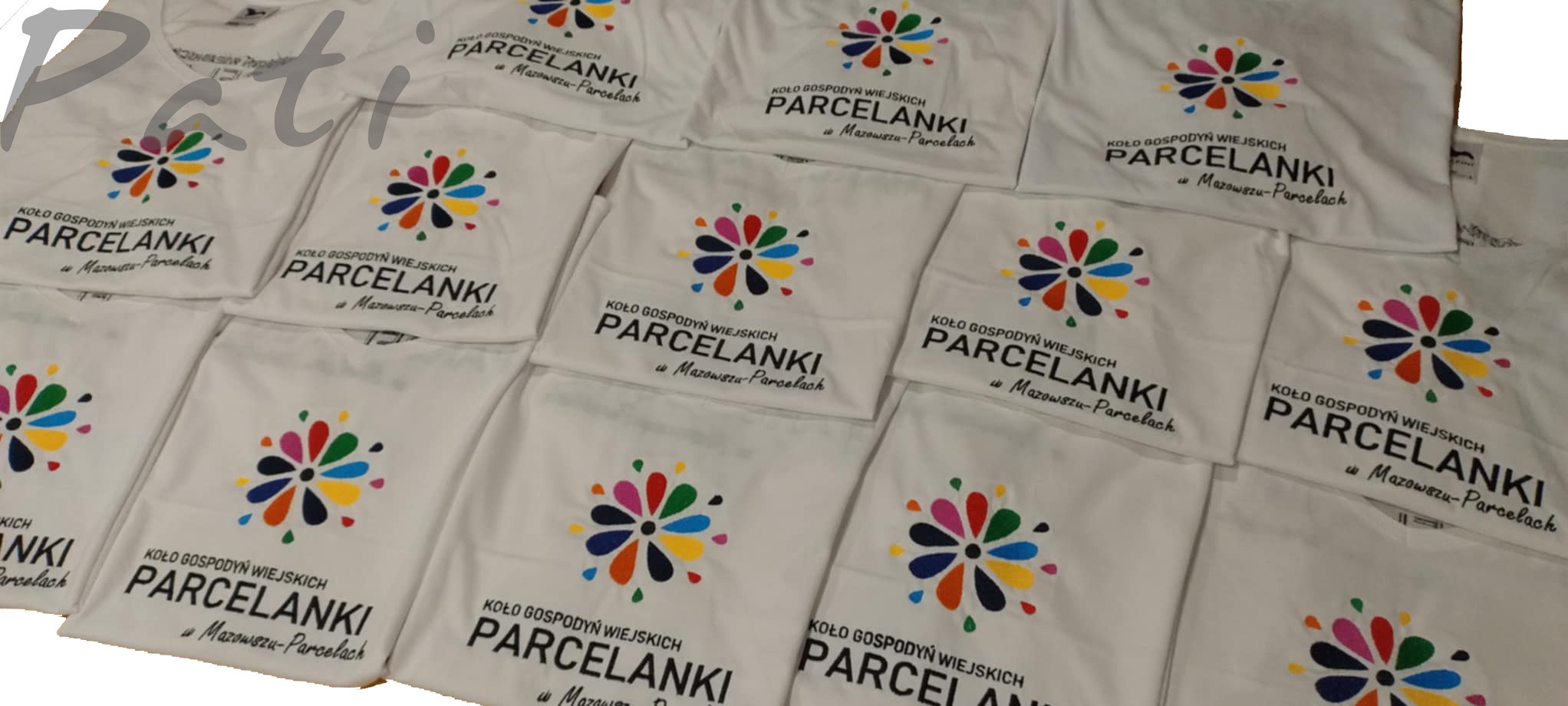 Białe koszulki z nadrukiem 'Koło Gospodyń Wiejskich Parcelanki' i kolorowym kwiatem, ułożone warstwami. Widoczne detale nadruku i materiału.