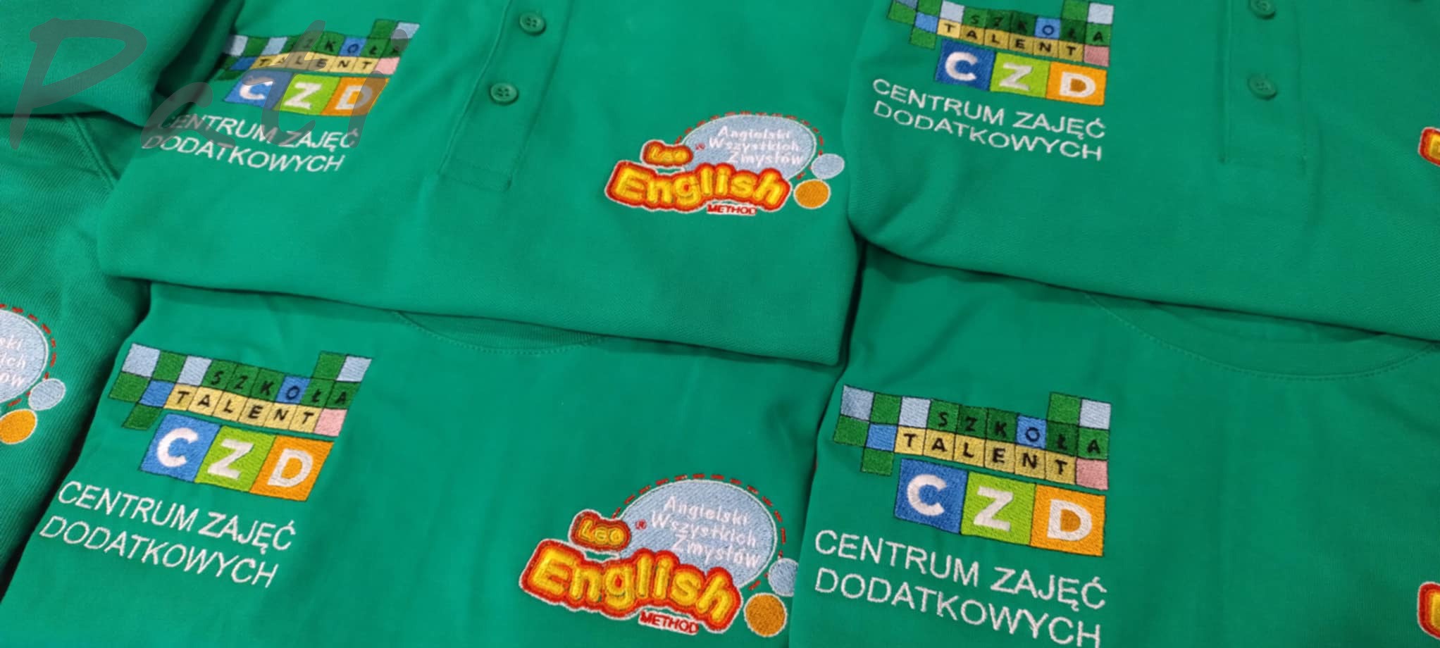 Zielone koszulki polo z haftowanymi logotypami i napisami 'Szkoła Talent CZD Centrum Zajęć Dodatkowych' oraz 'Leo English'. Precyzyjne, kolorowe detale haftu.