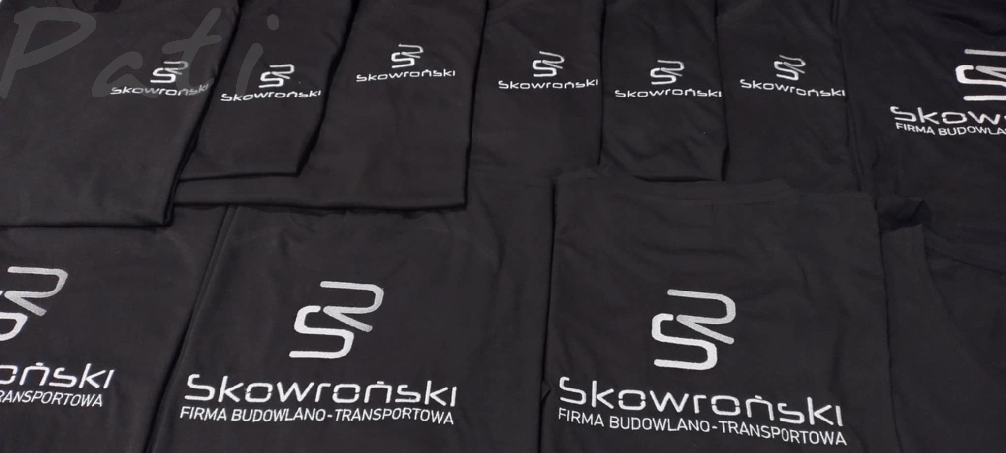 Czarne koszulki z białym nadrukiem logo firmy budowlano-transportowej, ułożone obok siebie. Widoczne detale nadruku i materiału.