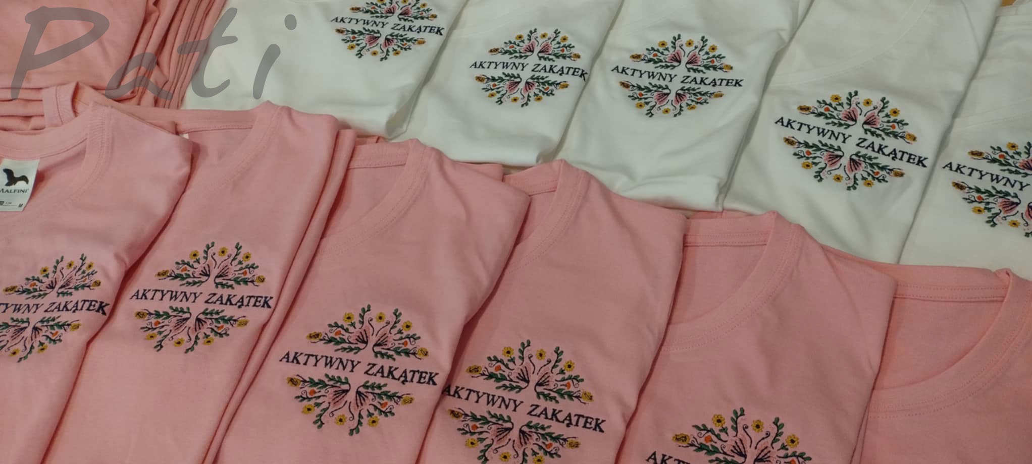 Stos różowych i białych koszulek z haftowanym motywem kwiatowym i napisem 'Aktywny Zakątek'. Różne kolory t-shirtów, zbliżenie na detale haftu.