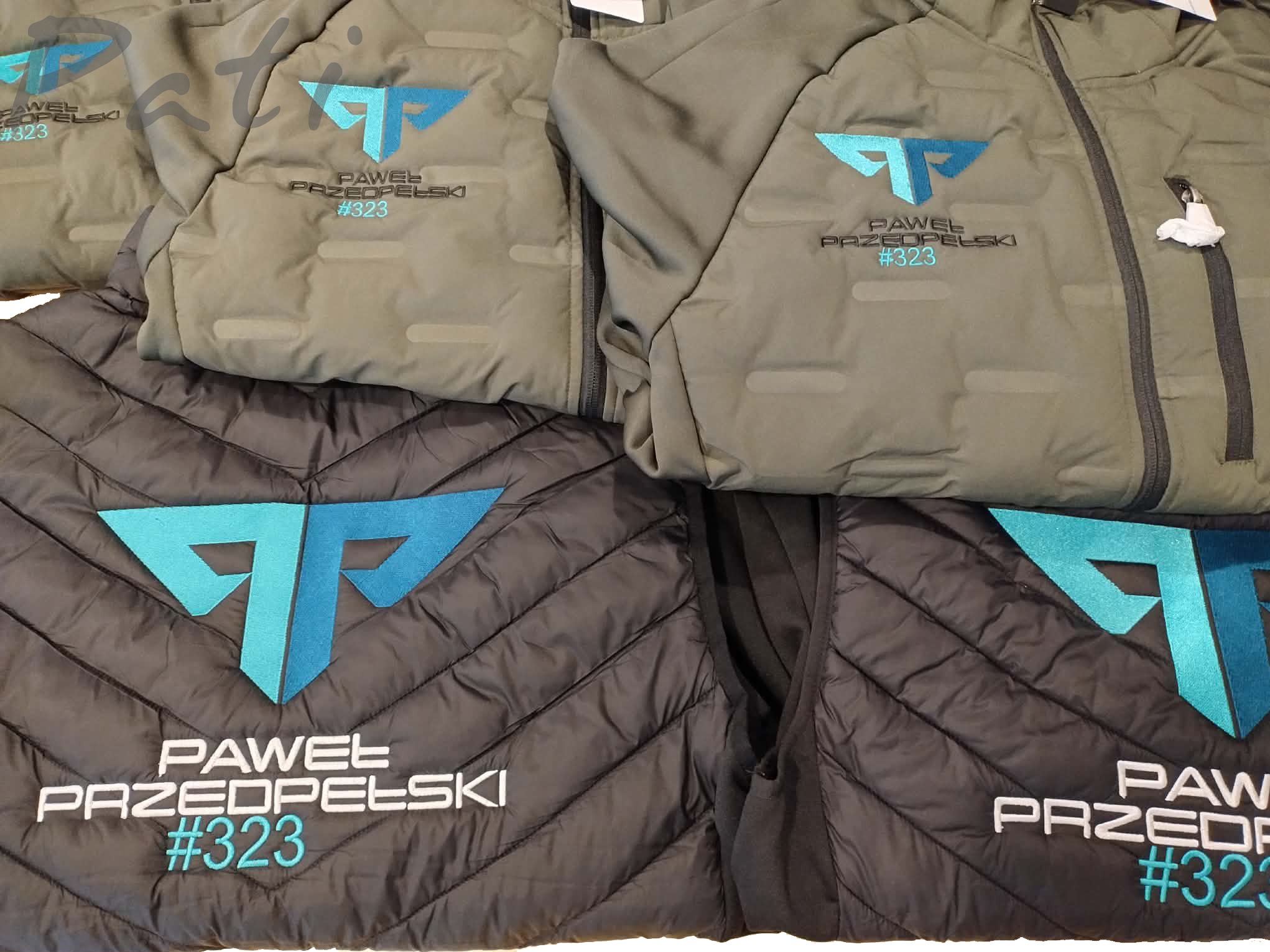 Kurtki i kamizelki z haftowanym logo i nazwą 'Paweł Przedpełski #323' w odcieniach szarości i khaki. Widoczne detale zamków i pikowania.