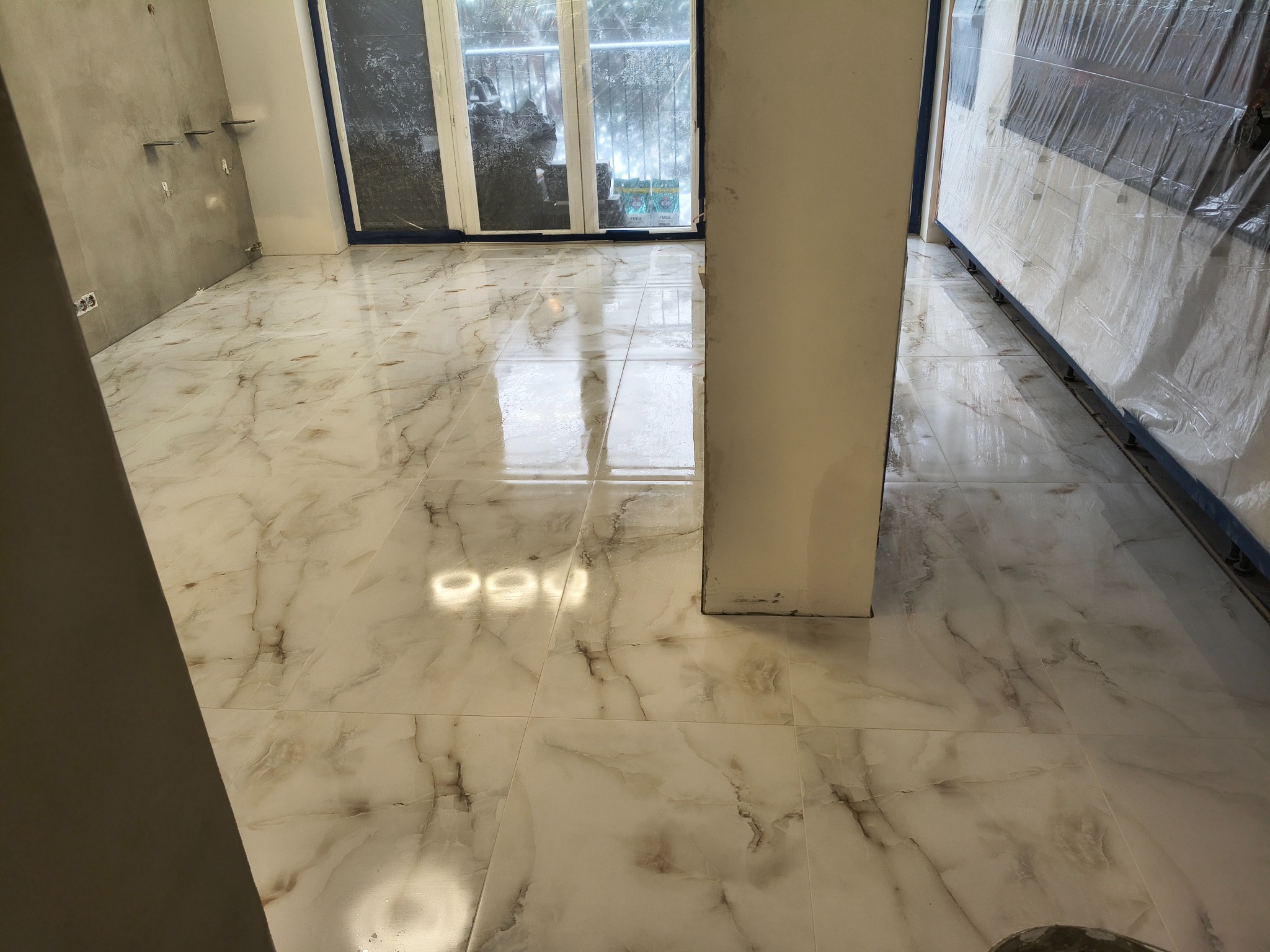 Połyskująca, marmurowa podłoga w jasnych odcieniach, świeżo po ułożeniu. Widoczne surowe ściany i okno zabezpieczone folią malarską. Wnętrze w trakcie wykańczania.