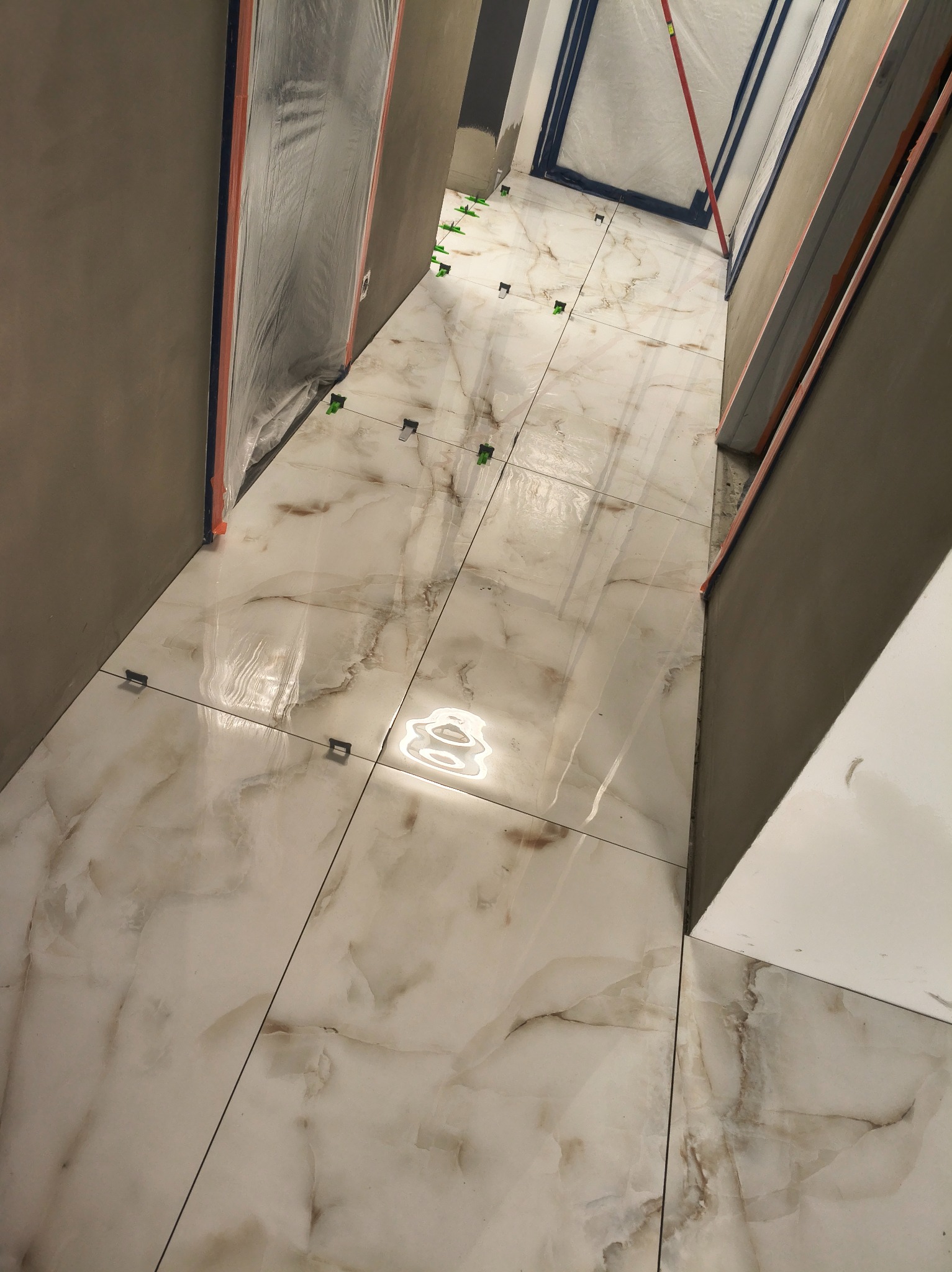 Układanie płytek imitujących marmur na podłodze w wąskim pomieszczeniu. Widoczne klipsy poziomujące. Ściany przygotowane do dalszych prac wykończeniowych.