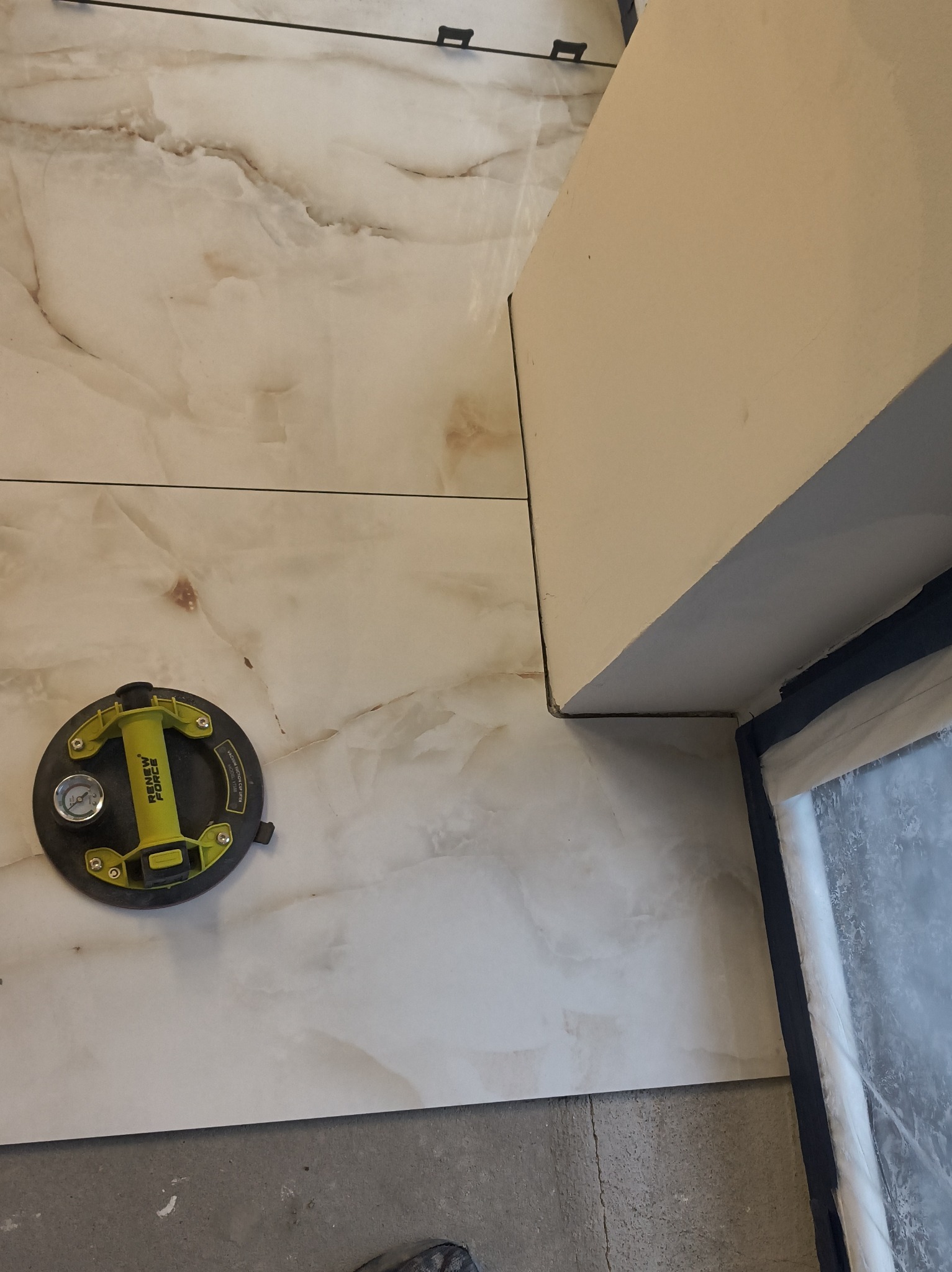 Ułożone płytki imitujące marmur, z przyssawką do płytek i elementami dystansowymi. Widoczny fragment okna i ściany. Jasne, neutralne kolory.