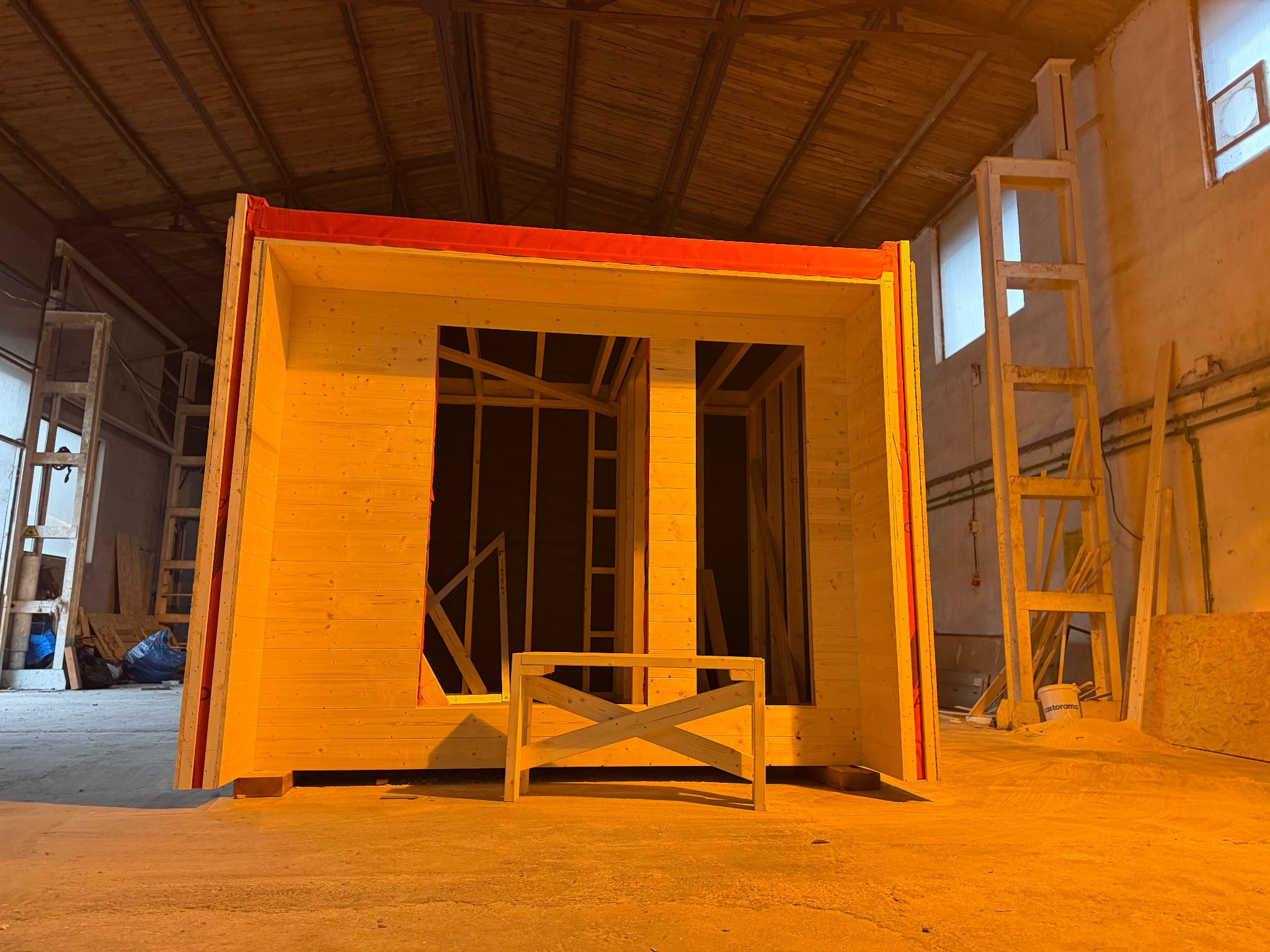 Wnętrze hali z surową konstrukcją drewnianą domu szkieletowego z czerwonymi elementami, widoczne deski i stelaż okienny, w tle drabina i materiały budowlane.