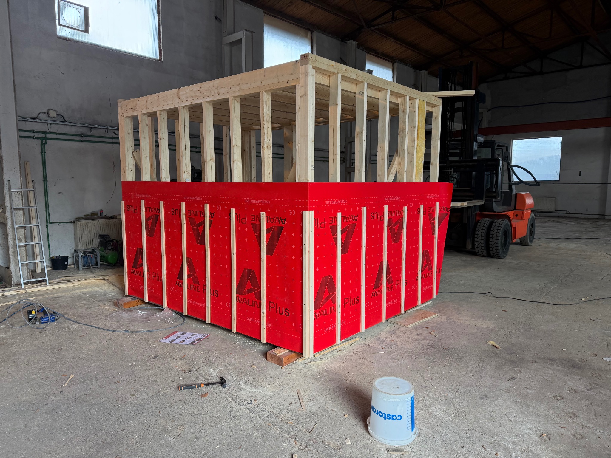 Wznoszenie szkieletu domu modułowego z czerwonymi panelami izolacyjnymi. Widoczny wózek widłowy w tle, narzędzia i wiadro farby na posadzce.