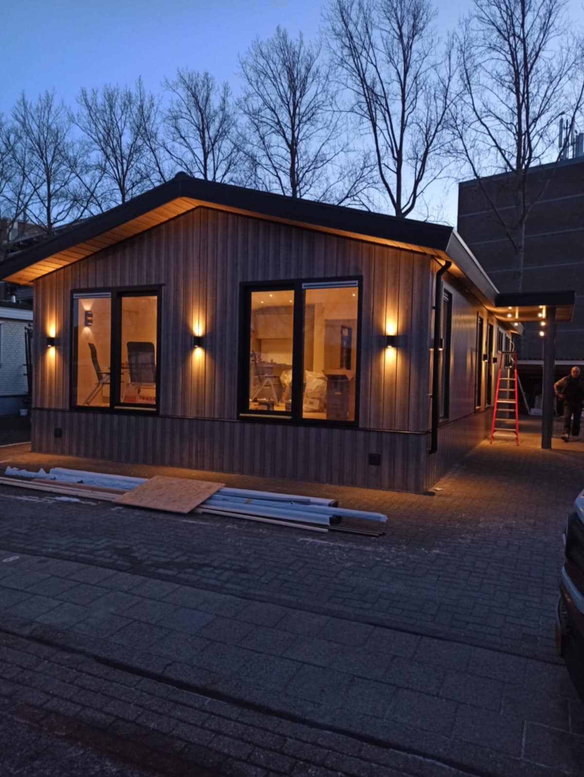 Nowoczesny dom modułowy z drewnianą elewacją oświetlony wieczornym światłem. Widoczne okna, schody i materiały budowlane na zewnątrz.