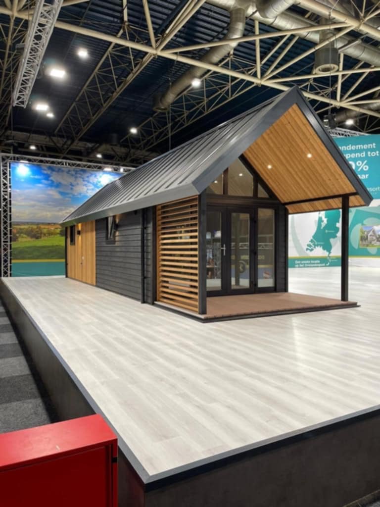 Nowoczesny, parterowy dom z szarego drewna z elementami z jasnego drewna i metalowym dachem na jasnej podłodze, prezentowany na tle ekspozycji targowej.