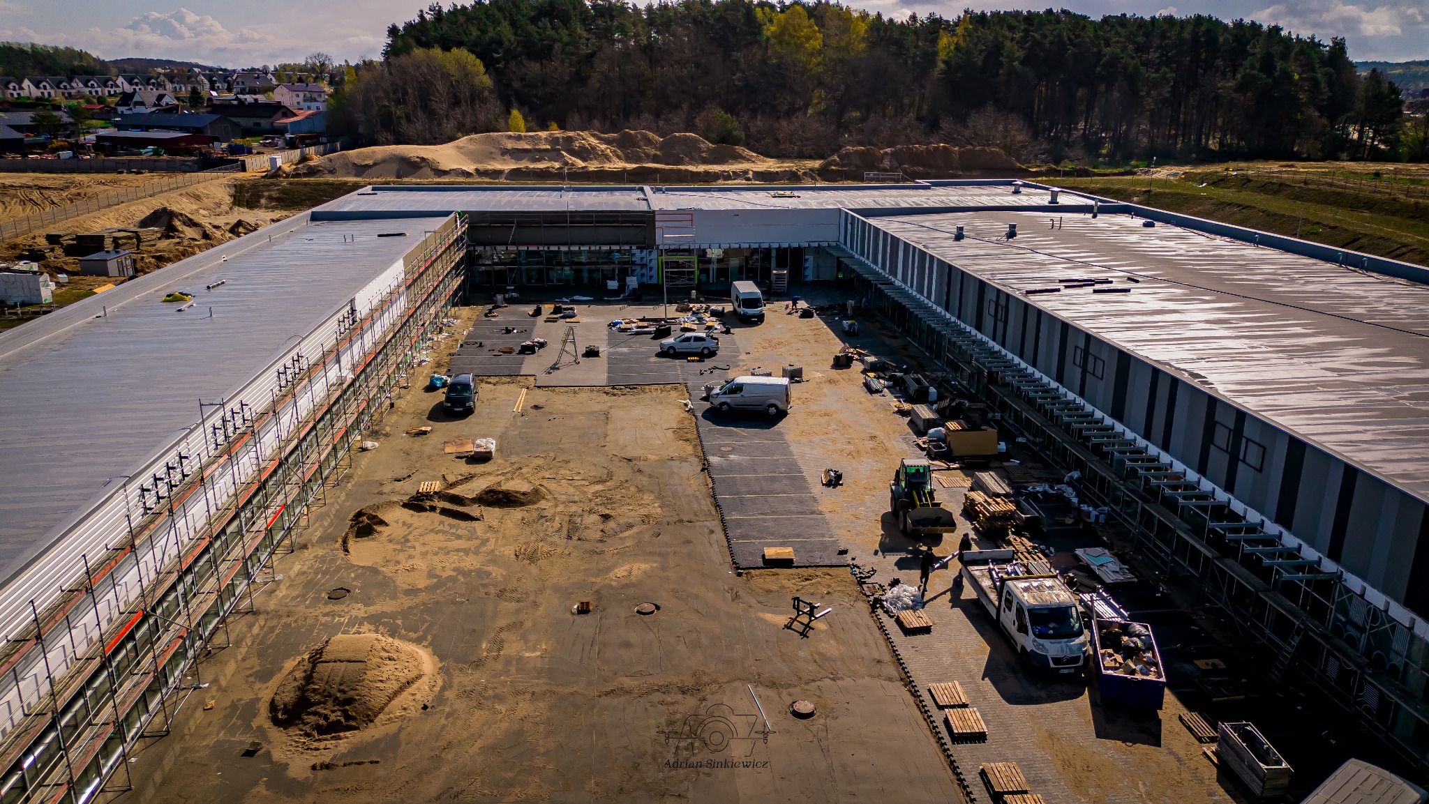 Rozległy budynek komercyjny w trakcie budowy, widok z lotu ptaka. Widoczne rusztowania, sprzęt budowlany i materiały. W tle zalesione wzgórza i zabudowa mieszkalna.