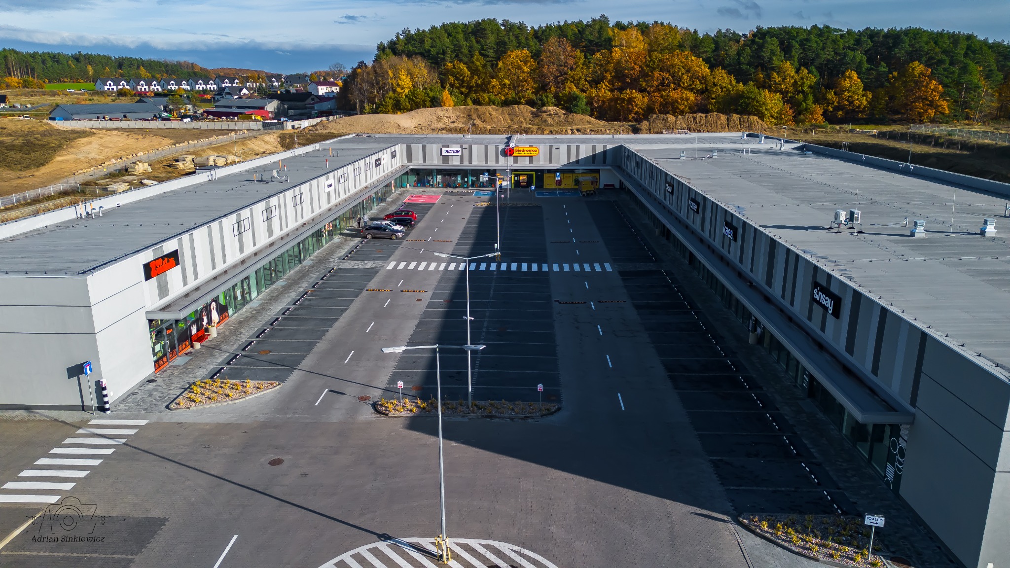 Widok z lotu ptaka na nowoczesne centrum handlowe z parkingiem, otoczone zielenią i zabudową mieszkalną w tle, w słoneczny dzień.