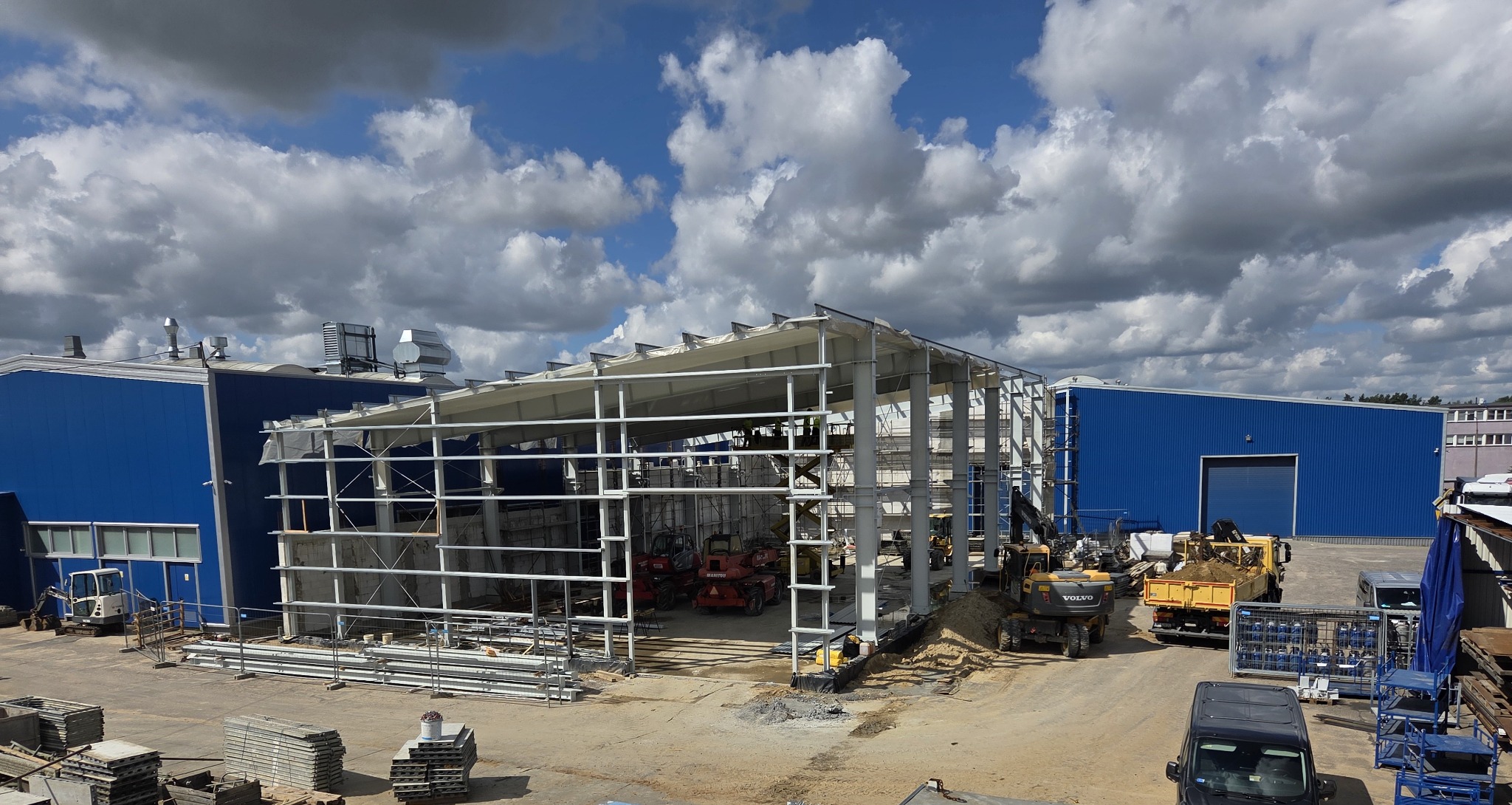 Konstrukcja stalowa hali w trakcie montażu, widoczne maszyny budowlane i błękitne budynki w tle, pochmurne niebo. Etap budowy z użyciem ciężkiego sprzętu.