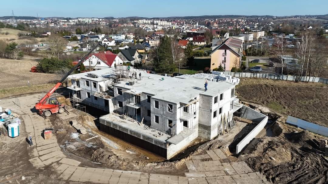 Wysoki kąt na budowę dwupiętrowego budynku z żelbetu w stanie surowym otwartym, z dźwigiem i pracownikami na dachu, na tle zabudowy miejskiej.