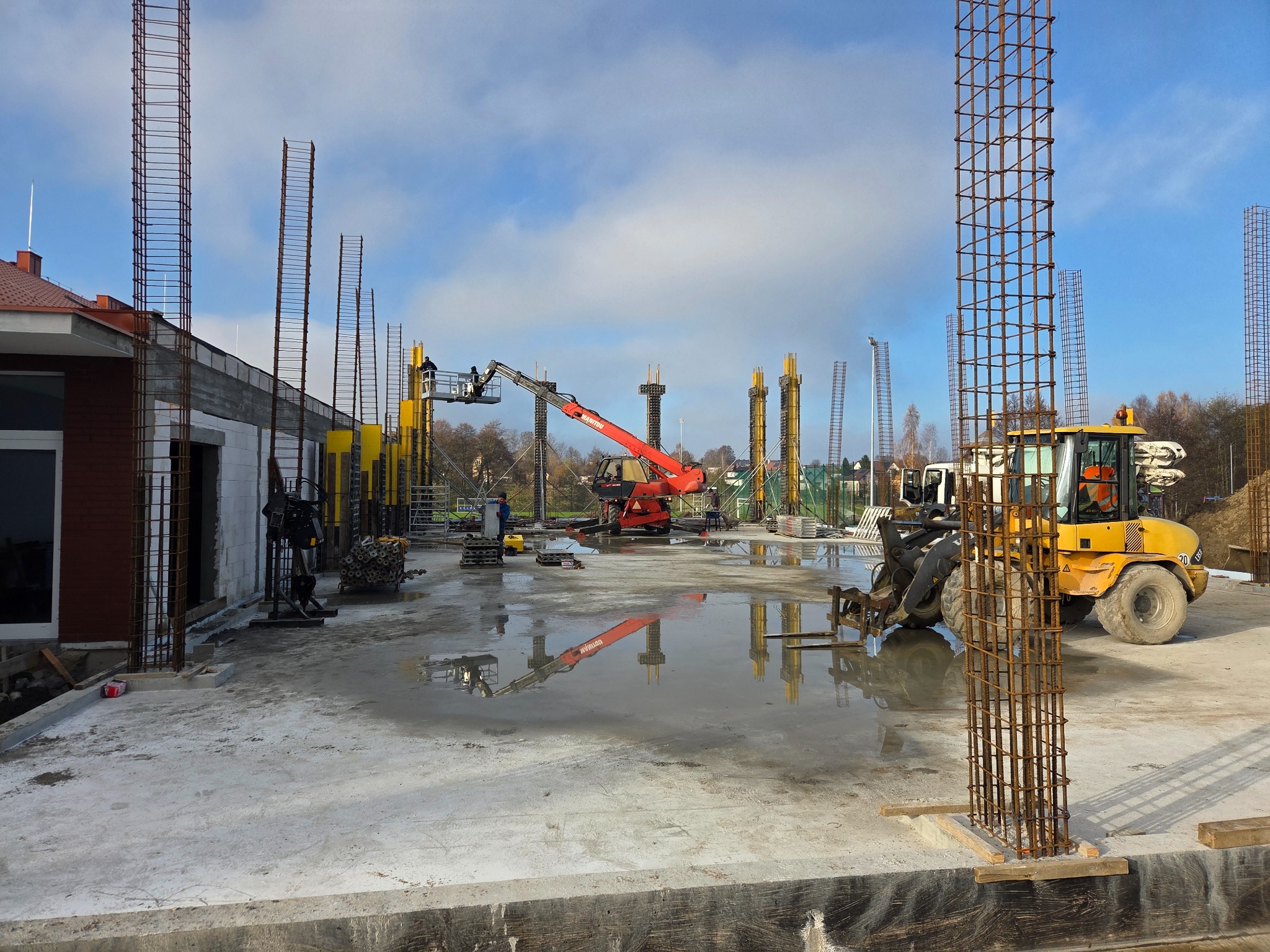 Wylewka fundamentowa z wystającymi zbrojeniami kolumn na budowie domu jednorodzinnego. W tle widoczny dźwig i maszyny budowlane.