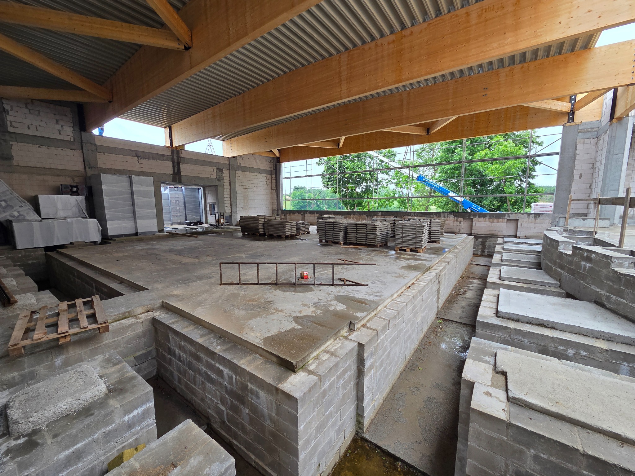 Wnętrze budynku w trakcie budowy z widocznymi fundamentami, ścianami z bloczków betonowych i drewnianą konstrukcją dachu. Na zewnątrz widoczny dźwig.