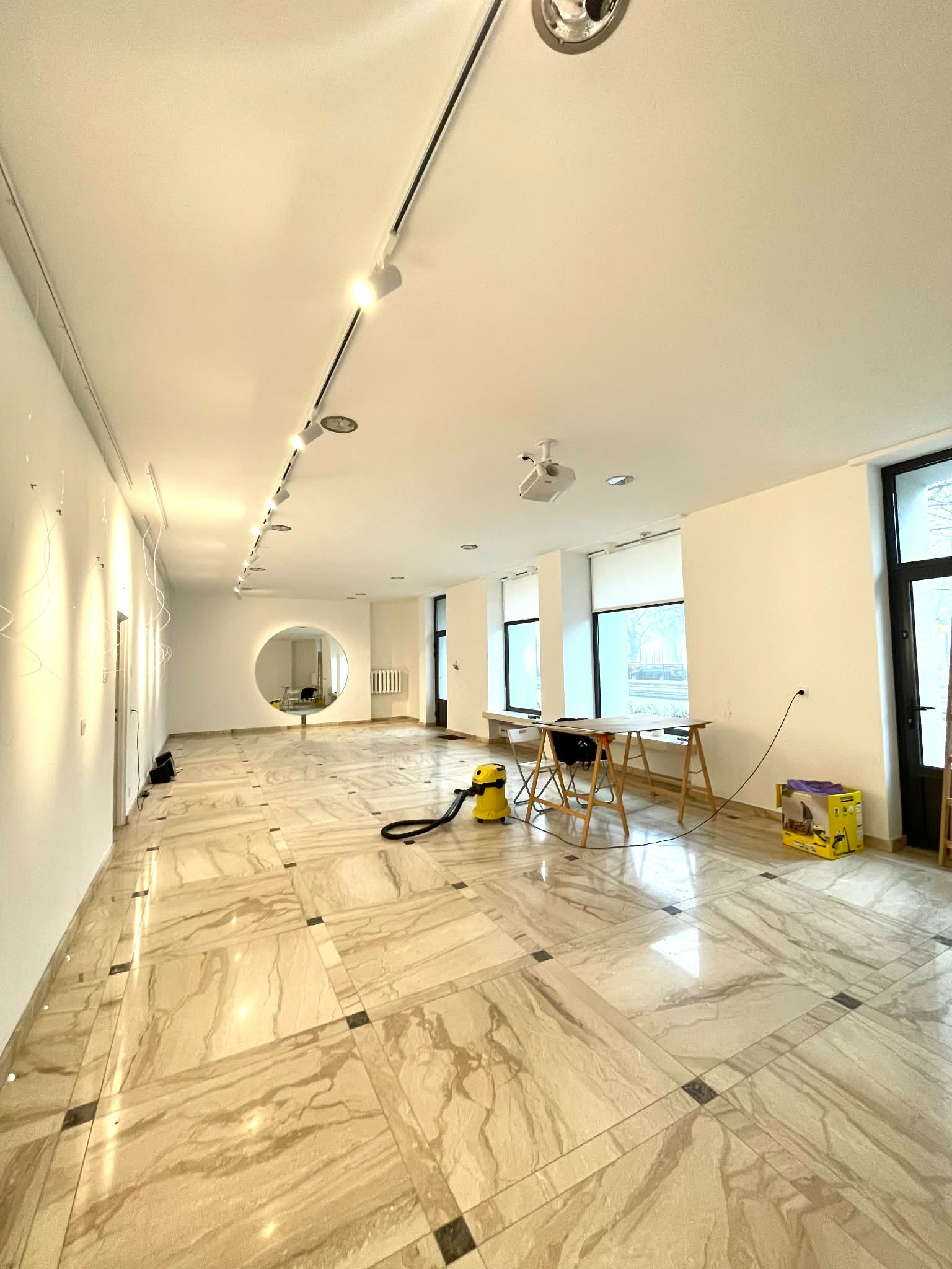 Jasne, przestronne wnętrze z marmurową podłogą w kwadraty, oświetleniem szynowym i okrągłym oknem. Widoczne narzędzia i drabina sugerują prace wykończeniowe.