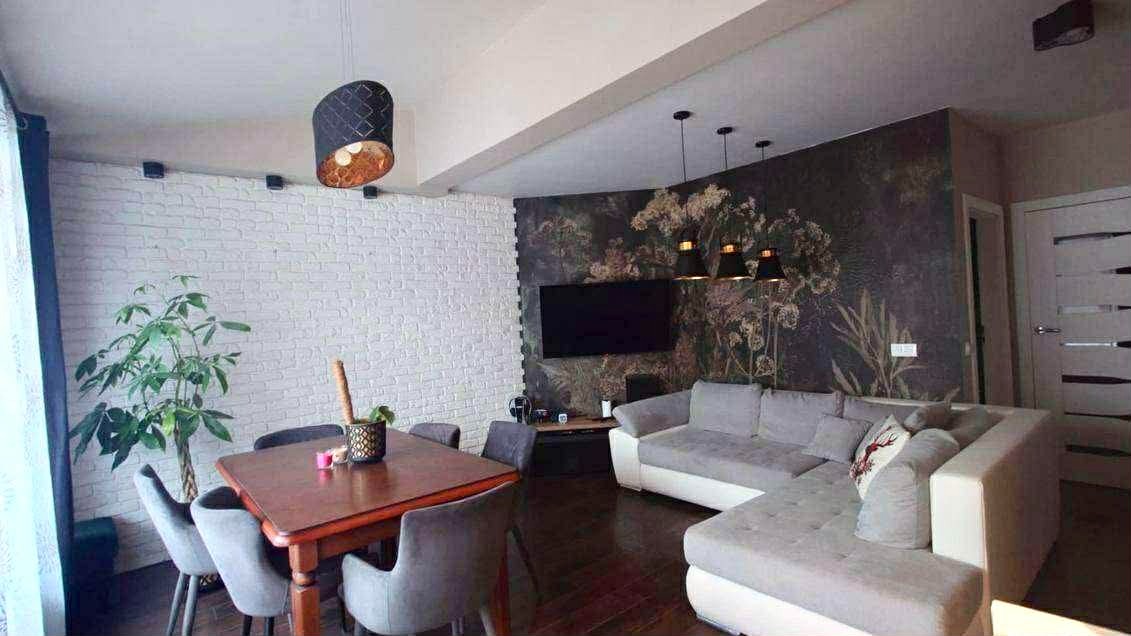 Nowoczesny salon z białą cegłą, tapetą w kwiaty i szarą sofą narożną. Widoczny stół z krzesłami i designerska lampa wisząca. Całość tworzy spójną aranżację wnętrza.