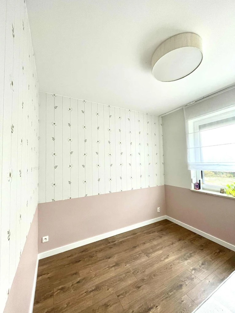 Pokój po odświeżeniu: ściany z tapetą w kwiatki i różową farbą do połowy, drewniana podłoga, okno z roletą i lampa sufitowa. Estetyczne wykończenie wnętrza.