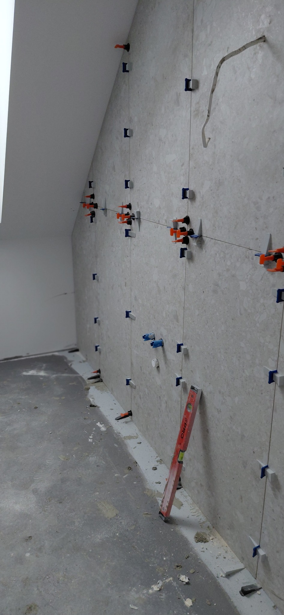 Ściana w trakcie układania płytek, widoczne systemy poziomowania i poziomica. Płytki w odcieniach szarości z delikatnym wzorem. Ukośny sufit w tle.