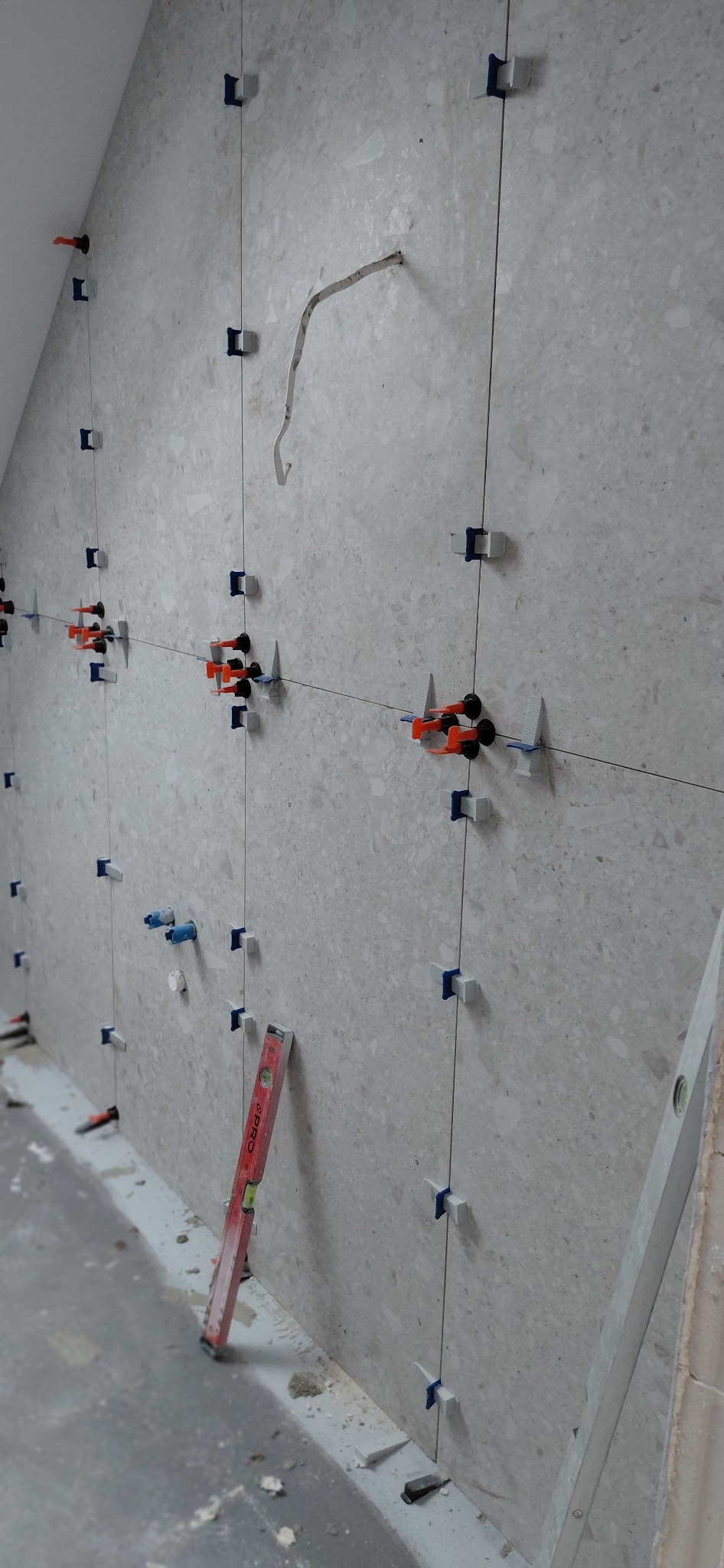 Ściana w trakcie układania płytek, z użyciem poziomicy i systemu poziomowania. Widoczne elementy mocujące i sznurki wyznaczające linie. Jasne płytki o nieregularnej fakturze.