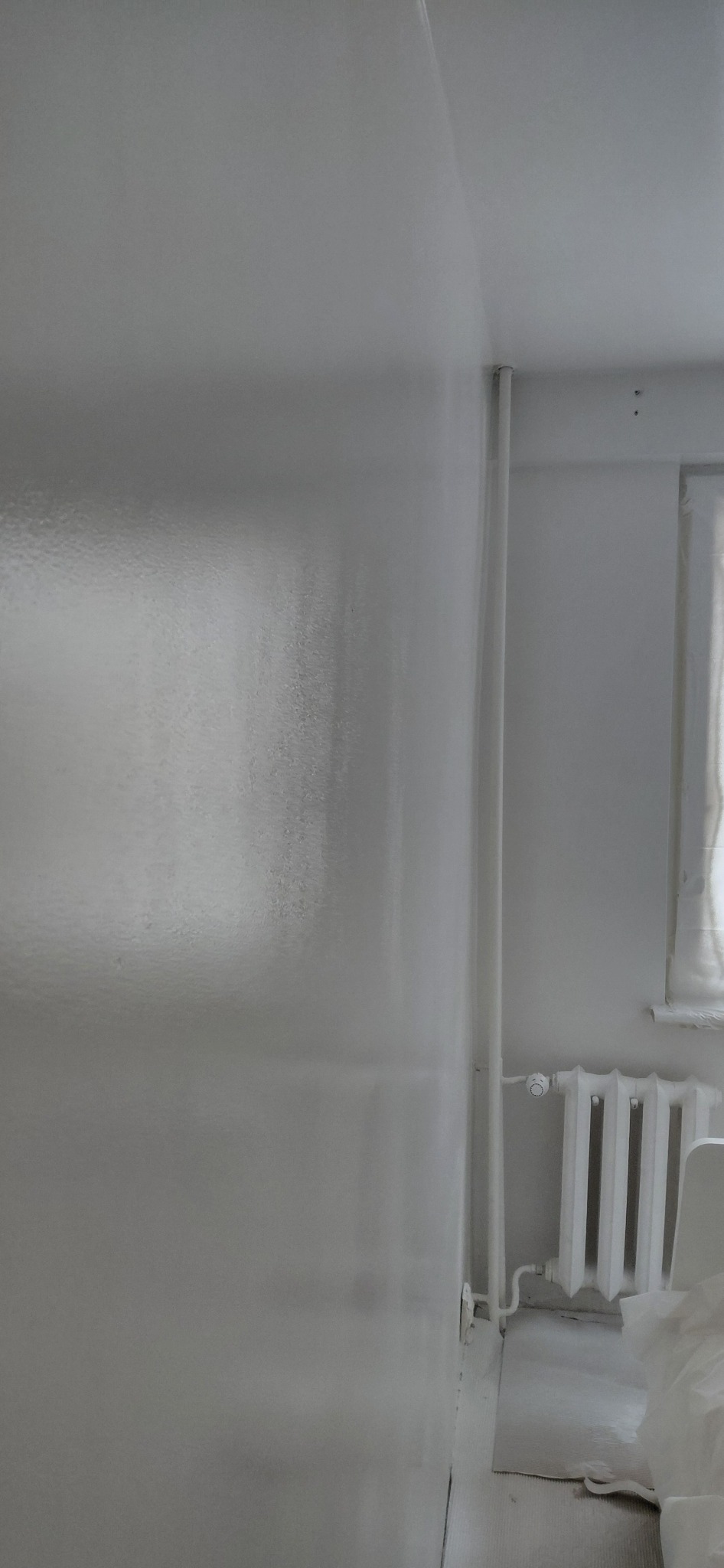 Biała ściana po malowaniu, widoczne delikatne refleksy światła, grzejnik i fragment okna w tle. Minimalistyczne wnętrze po odświeżeniu.
