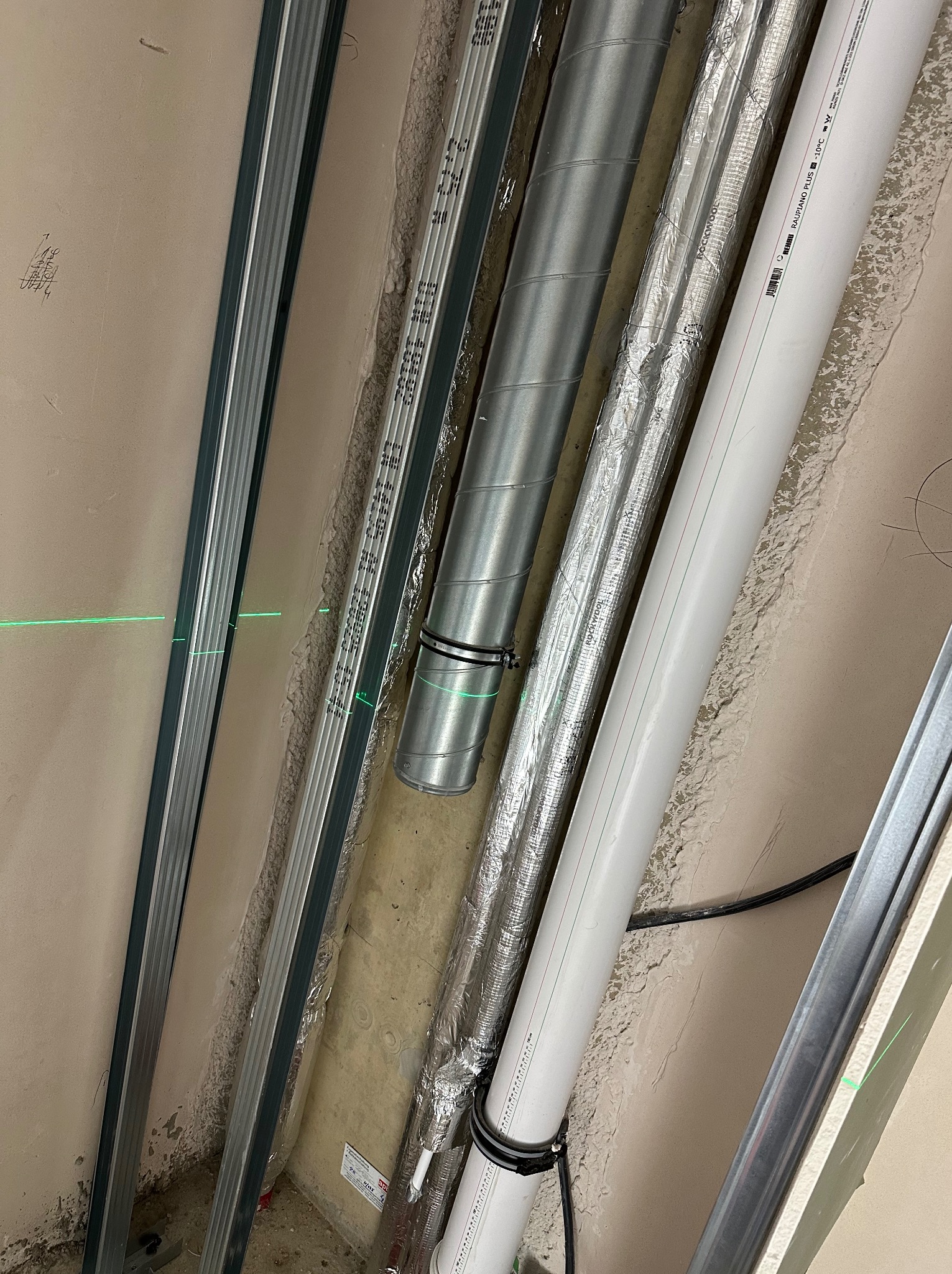 Instalacja rur wentylacyjnych i hydraulicznych w przestrzeni między ścianą a profilem, z widoczną zieloną linią lasera poziomującego.