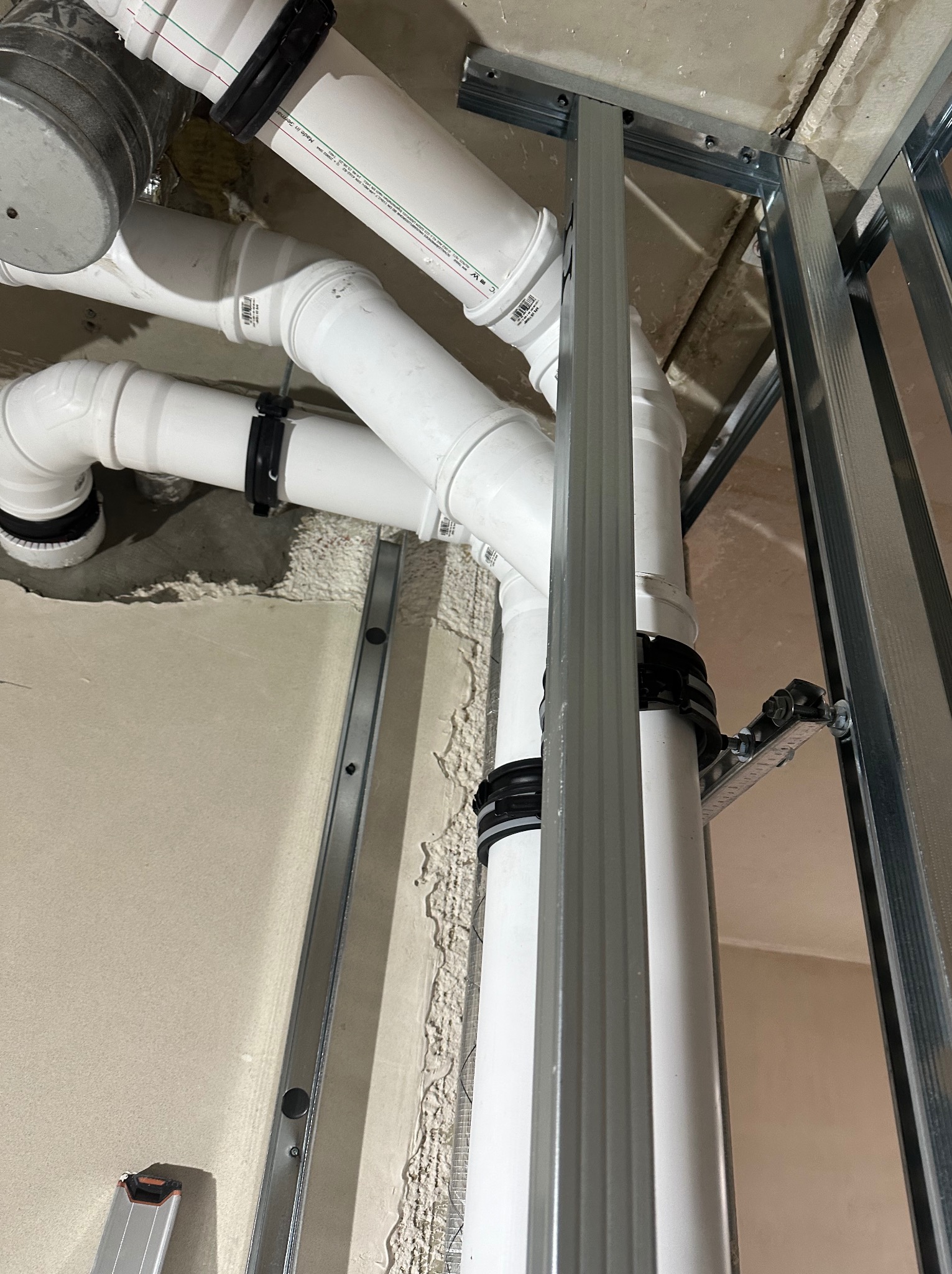 Białe rury kanalizacyjne z czarnymi obejmami, zamontowane pod sufitem, widoczne elementy konstrukcji metalowej. Instalacja w trakcie montażu.