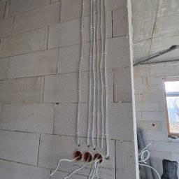 Instalacje Elektryczne Krystian Matła - Wewnętrzna instalacja elektryczna w surowym budynku: białe kable prowadzone wzdłuż ściany z betonu komórkowego, wchodzące do puszek instalacyjnych.