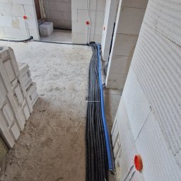 Instalacje Elektryczne Krystian Matła - Wnętrze w trakcie prac: wiązka przewodów w peszlach ułożona wzdłuż ściany z białego bloczka, widoczne puszki instalacyjne z przewodami. Surowy stan.