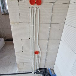 Instalacje Elektryczne Krystian Matła - Ściana z betonu komórkowego z zamontowanymi puszkami elektrycznymi i przewodami w białych peszlach. Widoczne również rury karbowane i niebieska rura.