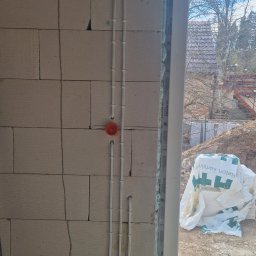 Instalacje Elektryczne Krystian Matła - Białe przewody elektryczne prowadzone pionowo po ścianie z betonu komórkowego, z pomarańczową puszką instalacyjną. Widok na okno i teren budowy w tle.
