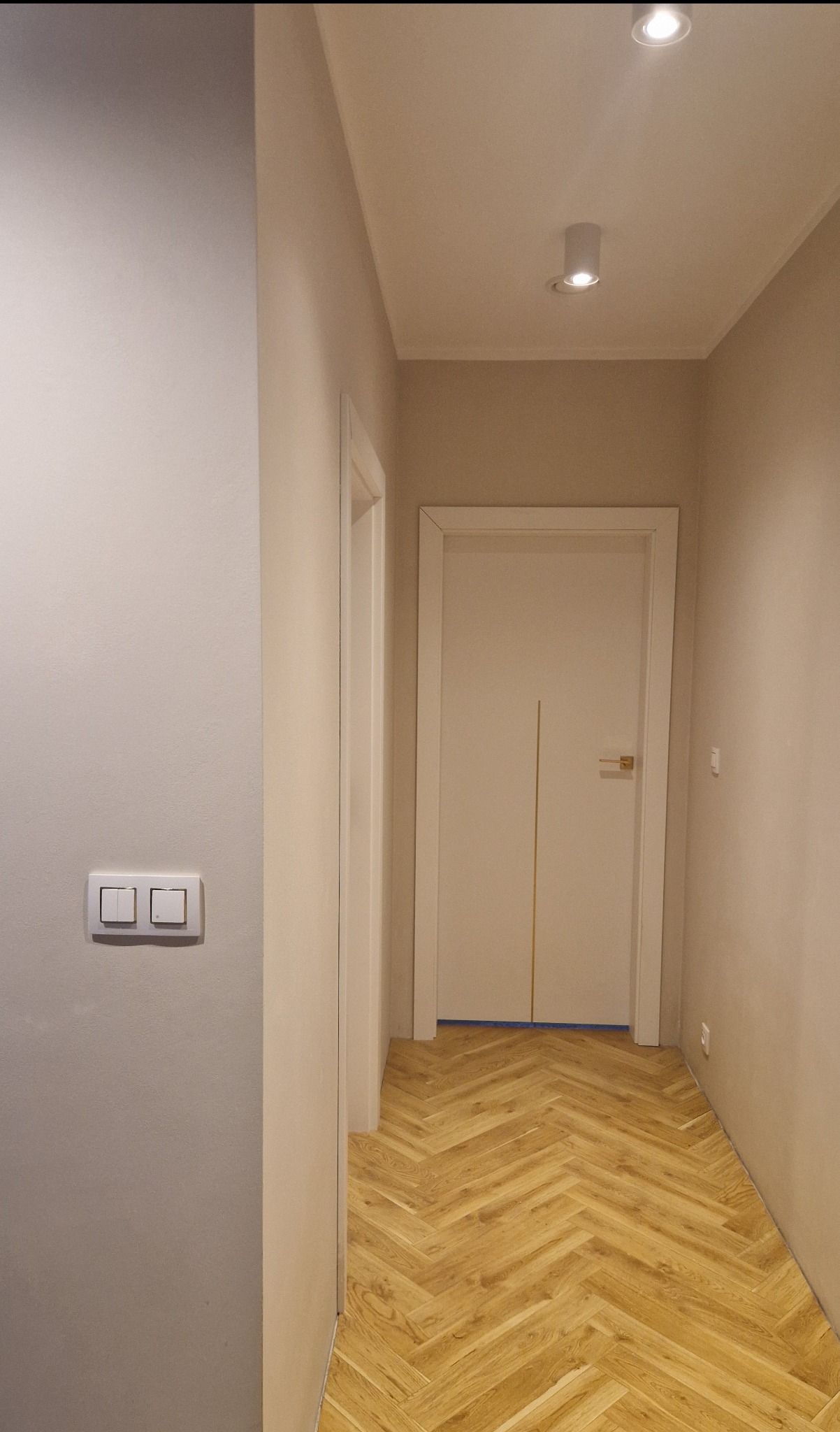 Korytarz w jasnych barwach z parkietem w jodełkę. Na ścianie widoczny podwójny włącznik światła. Oświetlenie sufitowe punktowe.