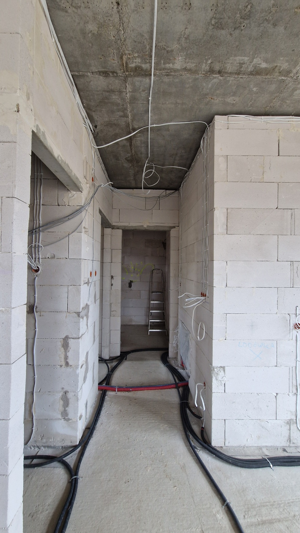 Surowy stan: Instalacja przewodów elektrycznych i rur w nowym budynku z białych bloków. Widoczne kable na ścianach i podłodze, drabina w tle.