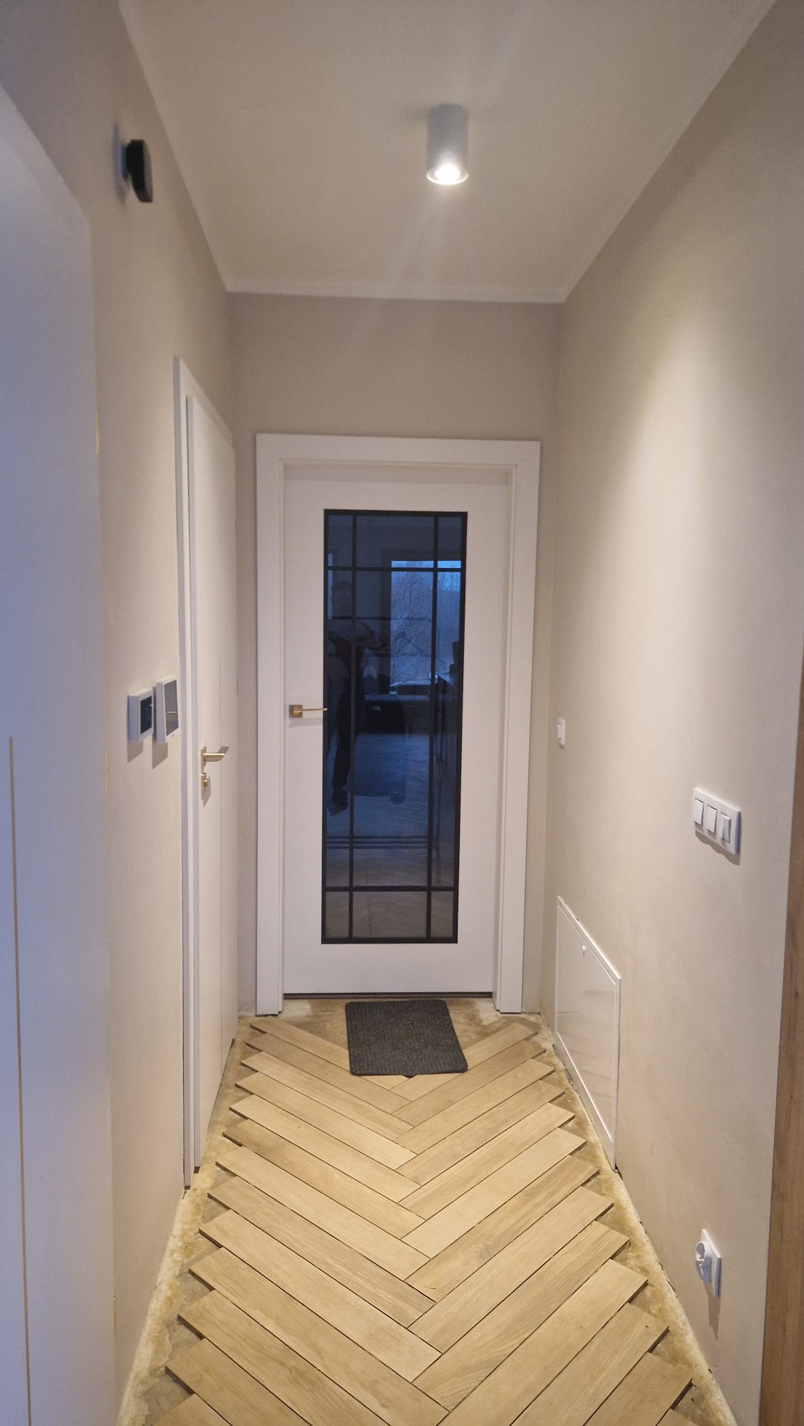 Korytarz z parkietem ułożonym w jodełkę, białe drzwi z szybą i włączniki światła na ścianie. Widoczna lampa sufitowa i czujnik. Neutralna kolorystyka ścian.