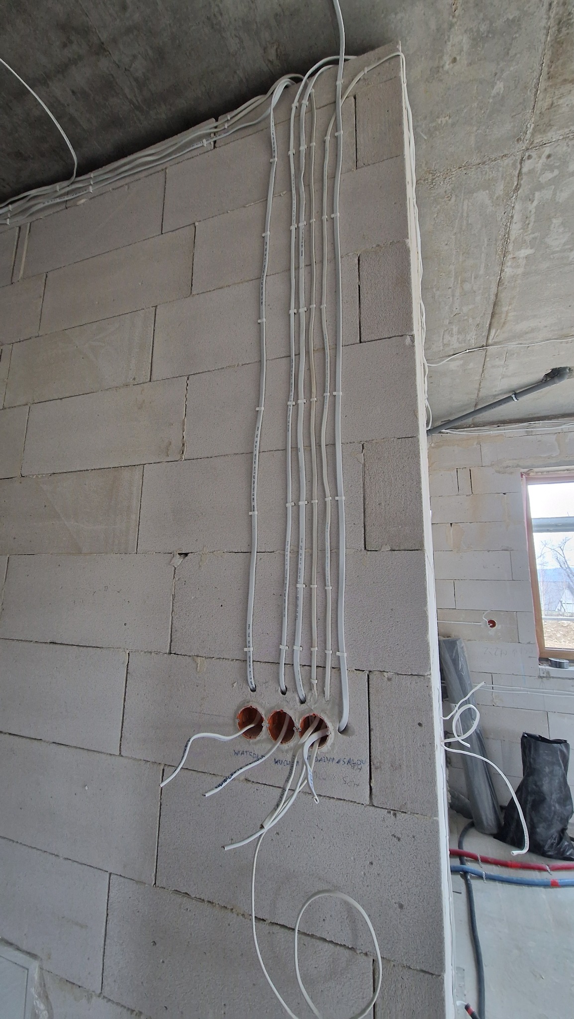 Wewnętrzna instalacja elektryczna w surowym budynku: białe kable prowadzone wzdłuż ściany z betonu komórkowego, wchodzące do puszek instalacyjnych.