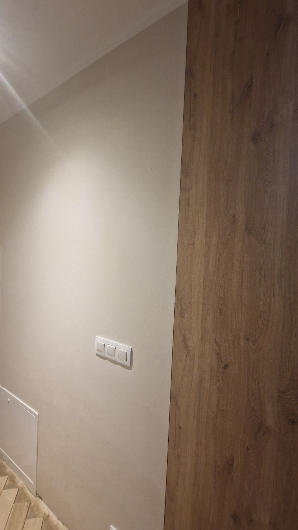 Ściana z włącznikami światła i panelem drewnopodobnym. Widoczne trzy białe włączniki na jasnej ścianie oraz fragment podłogi.