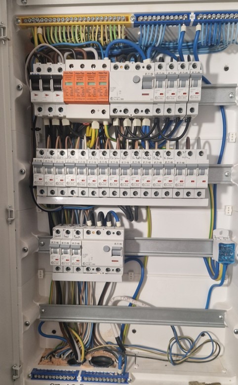 Rozdzielnia elektryczna w białej obudowie, widoczne zabezpieczenia, przewody w różnych kolorach, listwy zaciskowe. Ujęcie z bliska, detale instalacji.