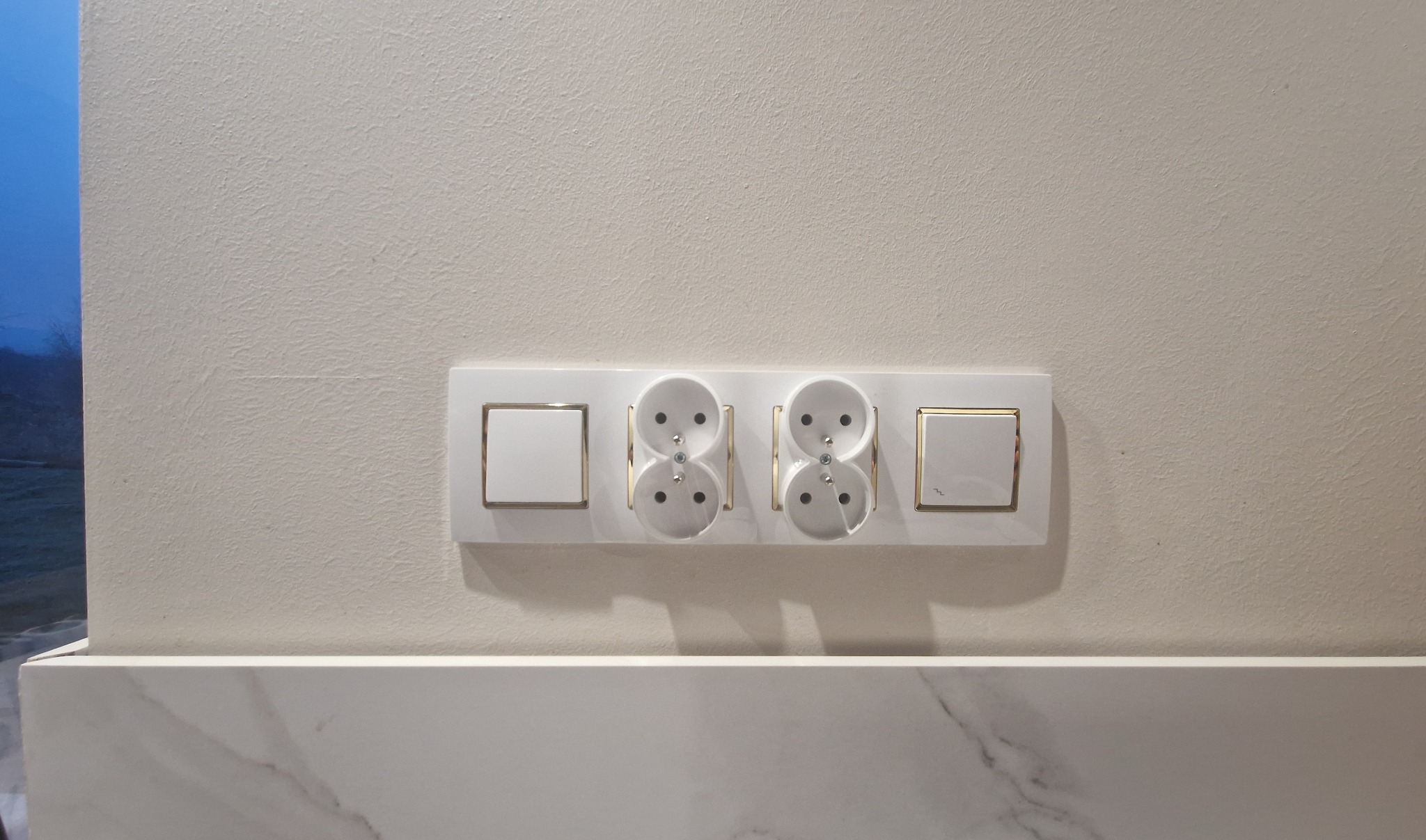 Biały panel z dwoma gniazdkami i włącznikami światła z delikatnym złotym obramowaniem, zamontowany na kremowej ścianie nad marmurowym blatem.