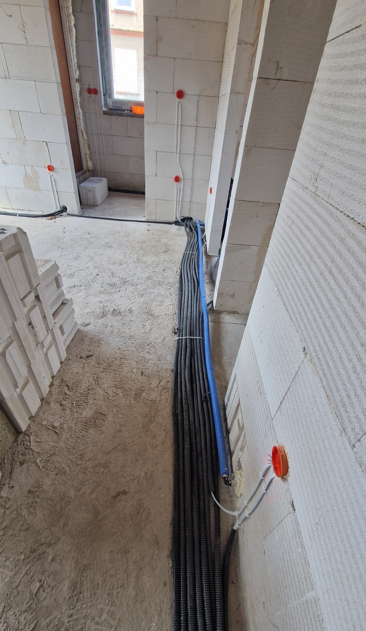Wnętrze w trakcie prac: wiązka przewodów w peszlach ułożona wzdłuż ściany z białego bloczka, widoczne puszki instalacyjne z przewodami. Surowy stan.