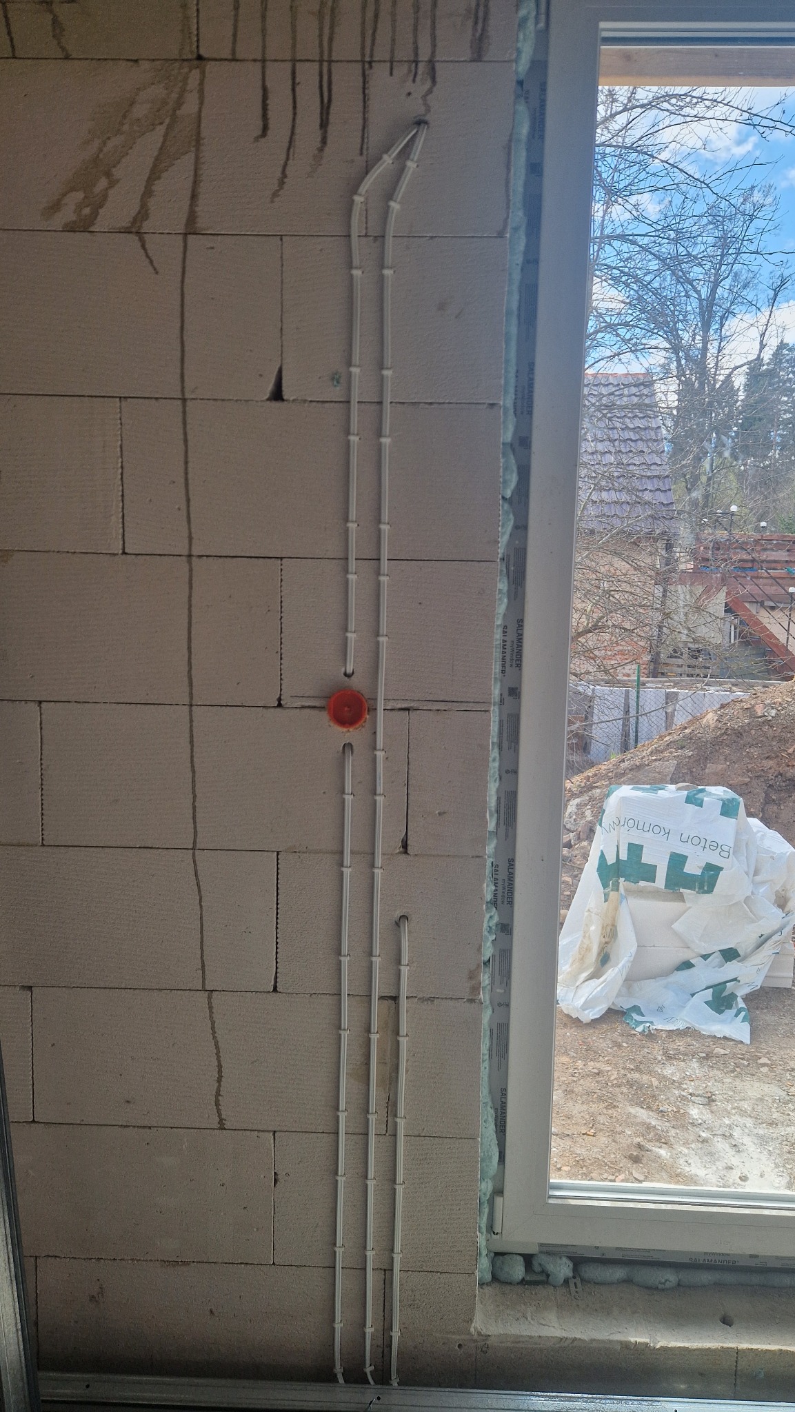 Białe przewody elektryczne prowadzone pionowo po ścianie z betonu komórkowego, z pomarańczową puszką instalacyjną. Widok na okno i teren budowy w tle.