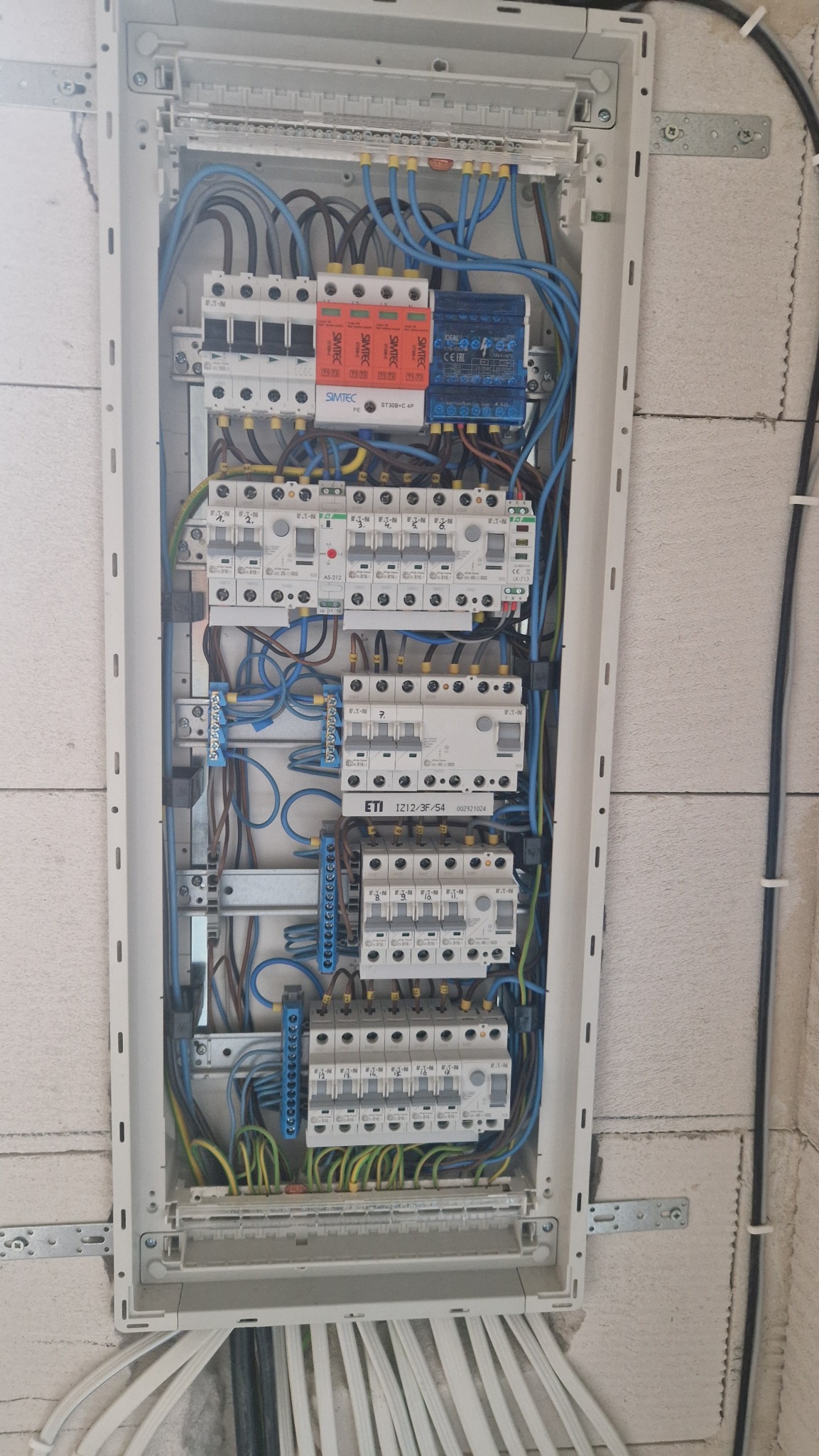 Rozdzielnica elektryczna z zainstalowanymi bezpiecznikami, okablowaniem w kolorach niebieskim, brązowym i żółto-zielonym, zamontowana na ścianie z betonu komórkowego.
