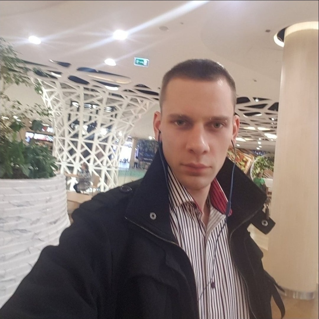 Mężczyzna w czarnej kurtce i koszuli w paski, z słuchawkami w uszach, na tle jasnego wnętrza z roślinami i kolumnami. Ujęcie z dołu.