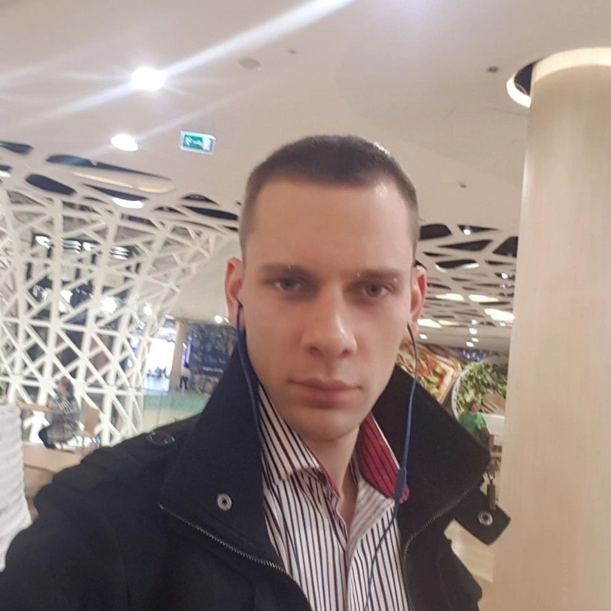 Mężczyzna w koszuli w paski i czarnej kurtce, z słuchawkami na szyi, na tle nowoczesnej konstrukcji sufitu w jasnym wnętrzu.