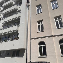 S.o.alpin.pro. - Pracownik na linie, w trakcie prac na elewacji budynku z jasną fasadą. Widoczne okna i elementy architektoniczne. U dołu zaparkowany samochód.