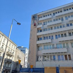 S.o.alpin.pro. - Dwóch alpinistów na linach odnawia elewację wysokiego budynku w mieście. Widoczne rusztowania i fragmenty odnowionej ściany. Błękitne niebo w tle.