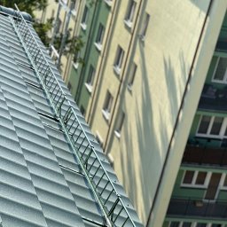 S.o.alpin.pro. - Szary dach z zamontowaną barierą przeciwśniegową na tle bloku mieszkalnego w słoneczny dzień. Widok z dołu, ukośny kadr.